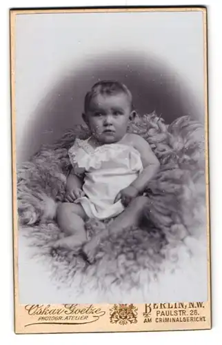 Fotografie Oskar Goetze, Berlin N. W., Paulsttr. 26, Baby mit zweifelndem Blick im weissen Gewand auf einem Pelz