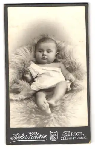 Fotografie Atelier Vicotria, Zürich, Limmat-Quai 22, Baby im weissen Gewand mit abwartendem Blick auf einem Pelz