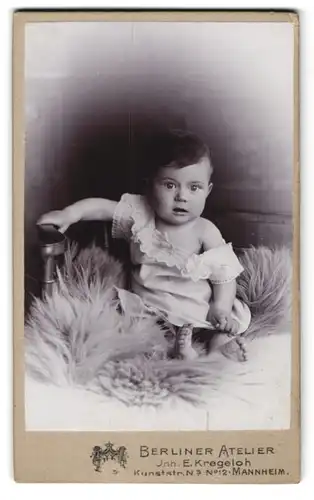 Fotografie Berliner Atelier, Mannheim, Kunststr. 12, Baby mit geschocktem Blick im weissen Gewand auf einem Pelz
