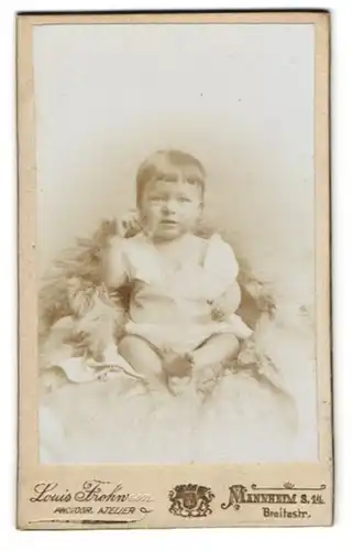 Fotografie Louis Frohwein, Mannheim, Breitestr, Kleinkind mit vollem Haar und weissem Kleid, auf einem Pelz sitzend