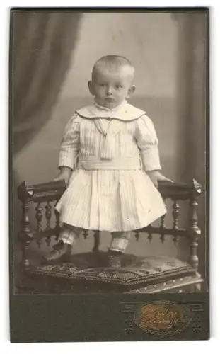 Fotografie E. Schuffert, Leipzig-Borna, Bahnhofstrasse 37, Kleinkind mit einem längsgestreiften Kleid auf einem Stuhl