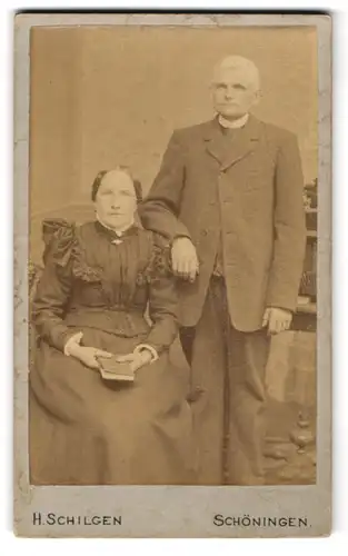 Fotografie H. Schilgen, Schöningen, Am Bahnhof, Altes bürgerliches Ehepaar, sie sitzend im Kleid mit verzierter Schulter