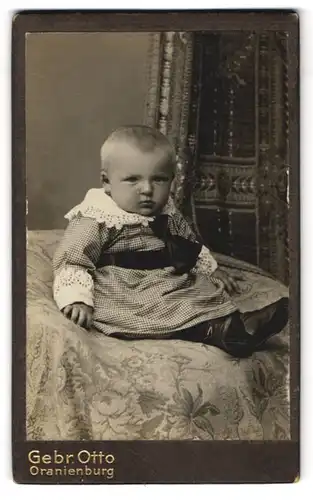 Fotografie Gebr. Otto, Oranienburg, Pausbäckiges Kleinkind im feinkarierten Kleid mit schwarzer Schleife und Zierkragen