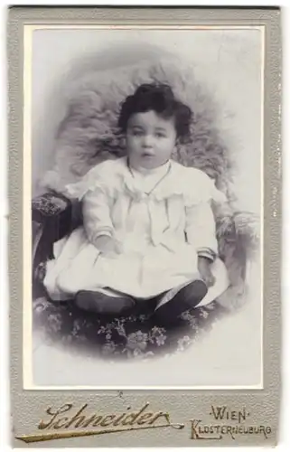 Fotografie Schneider, Wien, VII. Neubaugasse 29, Kleinkind mit schwarzem lockigen Haar und weissem Kleid auf einem Pelz