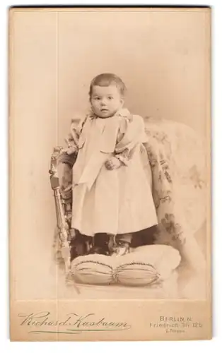 Fotografie Richard Kasbaum, Berlin N., Friedrich-Str. 125, Kleindkind im langen, weiten Kleid auf einem kissen stehend