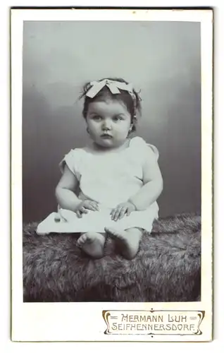 Fotografie Hermann Luh, Seifhennersdorf, Kleinkind mit Haarschleife in weissem Kleid sitzt brav da