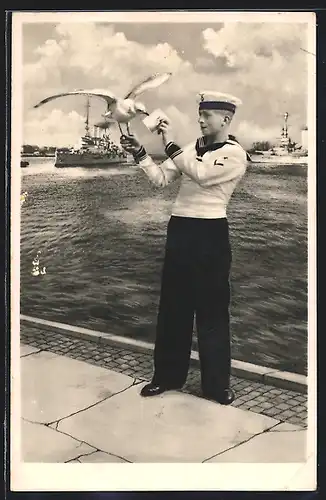 AK Eckernförde, Matrose mit Vogel auf der Hand, Im Hintergrund Schiff der Kriegsmarine
