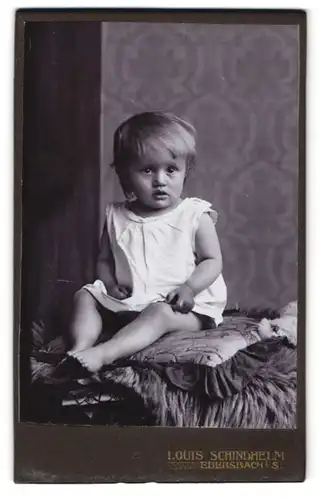 Fotografie Louis Schindhelm, Ebersbach i. S., Kleindkind im weissen Gewand mit fragendem Blick auf einem Sitzkissen