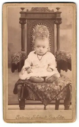 Fotografie Antonie Schmidt, Graslitz, Erstauntes Kleinkind im Rüschenkleid auf einem reich verzierten Stuhl
