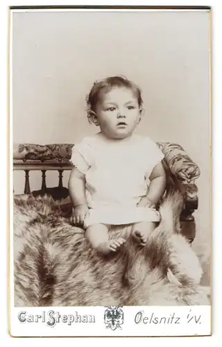 Fotografie Carl Stephan, Oelsnitz i. V., Kirchstr., Kleinkind in weissem Spitzenkleid sitzend auf einem Tierfell