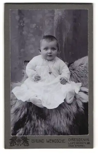 Fotografie Bruno Wendsche, Dresden, Leipzigerstr. 58, Niedliches Baby im weissen Kleid mit Herzkette auf Fell