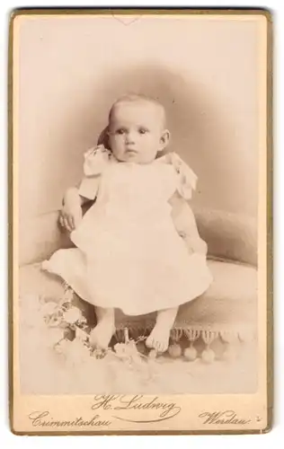 Fotografie H. Ludwig, Crimmitschau, Lindenstr., Niedliches Baby im weissen Kleid sitzt entspannt auf einem Sofa