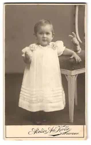 Fotografie Ad. Kolle, Göttingen, Prinzenstr. 18, Niedliches Kind im weissen Kleid mit runden Bäckchen fröhlich am Lächeln
