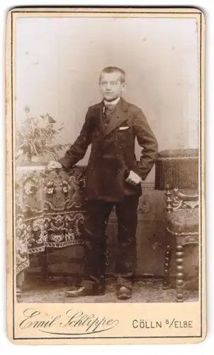 Fotografie Emil Schlippe, Cölln a. Elbe, Junger Bursche im schwarzen Anzug mit Krawatte nebst Tisch