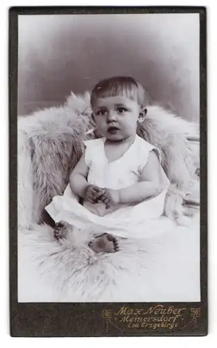 Fotografie Max Neuber, Meinersdorf i. Erzg., Kleindkind im weissen Gewand mit grossen Augen auf einem Pelz