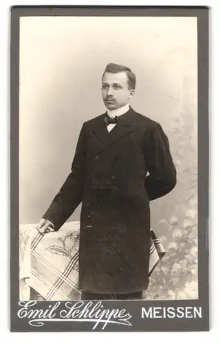 Fotografie Emil Schlippe, Meissen, Elbberg 1, Adretter junger Mann im schwarzen Mantel mit Fliege nebst Tisch