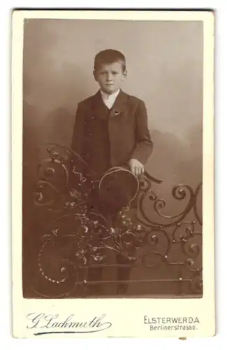 Fotografie G. Lachmuth, Elsterwerda, Berlinerstr., Junger Bursche im schwarzen Anzug mit Hut zur Hand und glattem Haar
