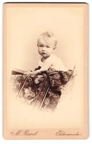 Fotografie M. Barol, Elsterwerda, Niedliches blondes Baby im weissen Kleid auf gemusteter Decke mit weit geöffnetem Mund