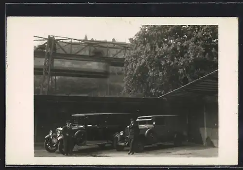 Foto-AK Auto Protos C1 10 /45 (1926), Zwei Chauffeure an ihrem geräumigen Fahrzeugen vor fahrender Schwebebahn