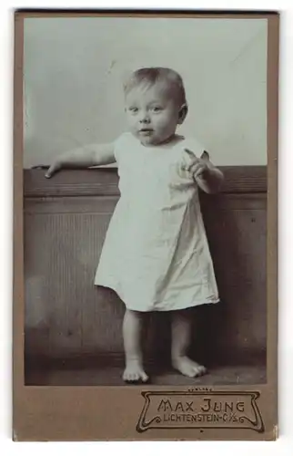 Fotografie Max Jung, Lichtenstein, Niedliches Kleinkind im weissen Kleid auf einer Bank zeigt nach vorne