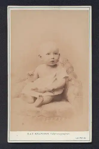 Fotografie A. & F. Naumann, Leipzig, Dorotheenstr. 6, Niedliches Baby im weissen Kleid mit erstauntem Blick und Glatze