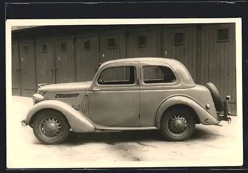 Foto-AK Auto Ford Eifel (1937), KFZ mit heller Karosserie und rückseitigem Ersatzreifen vor einer Reihe Garagen