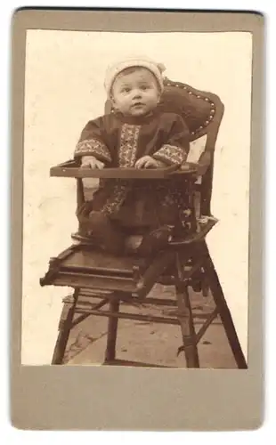Fotografie unbekannter Fotograf und Ort, Kleinkind im Kleid sitzt im Hochstuhl