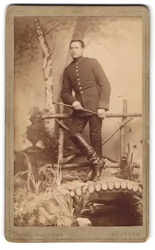 Fotografie Alex. Matthaey, Bautzen, Soldat in Uniform mit Gerte vor einer Studiokulisse