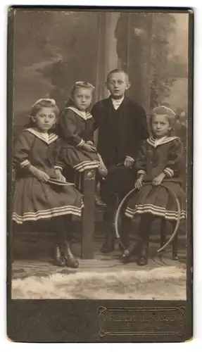 Fotografie Atelier Hertzsch, Leisnig, Drei Mädchen im identischen Kleid und ihr Bruder im schwarzen Anzug