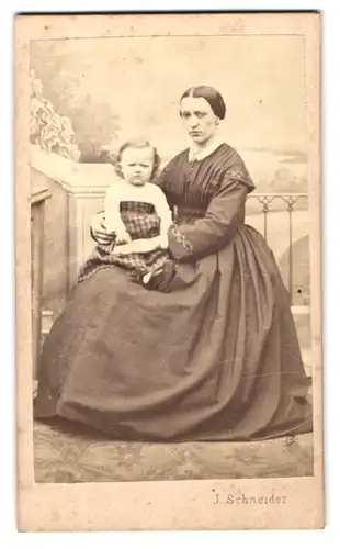 Fotografie Joh. Schneider, Wien, junge Mutter im dunklen Kleid mit ihrer Tochter auf dem Schoss vor einer Studiokulisse