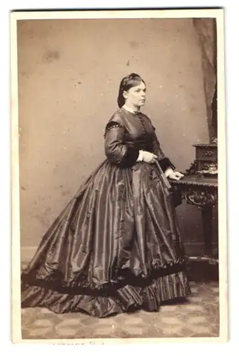 Fotografie W. Breuning, Hamburg, junge Dame im dunklen glänzenden Kleid steht am Sekreträr