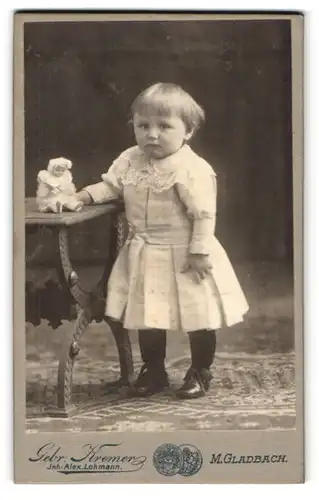 Fotografie Gebr. Kremer, Mönchen Gladbach, kleines Mädchen im weissen Kleid mit kleiner Puppe auf dem Tisch