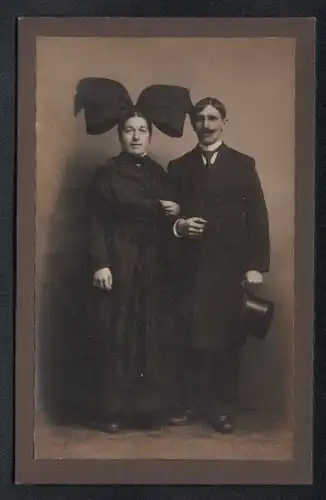 Fotografie unbekannter Fotograf und Ort, Frau im Trachtenkleid mit Schleife nebst Mann im Mantel mit Zylinder