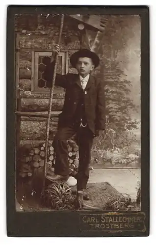 Fotografie Carl Stallechner, Trostberg, junger Knabe im Anzug mit Wanderstock vor einer Holzhütte, Studiokulisse