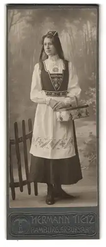 Fotografie Hermann Tietz, Hamburg, junge Frau in Tracht mit Sammelkorb vor einer Studiokulisse, 1906