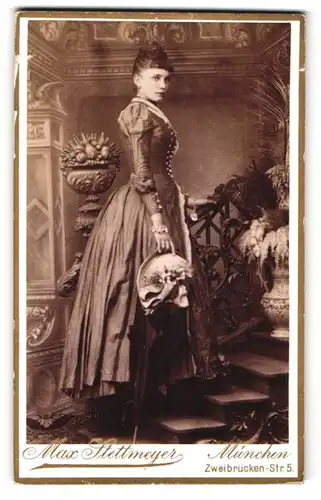 Fotografie Max Stettmeyer, München, hübsche junge Frau im edlen Kleid mit Hut auf einer Treppe, Studiokulisse