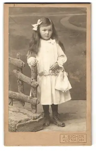 Fotografie A. Eicher, Uznach, niedliches kleines Mädchen im weissen Kleid mit offenen Haaren und Haarschleife