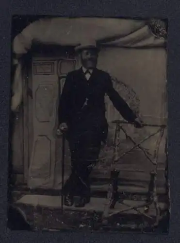 Fotografie Ferrotypie Herr im dunklen Anzug mit Stock und Hut vor einer Studiokulisse