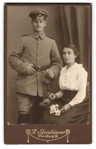 Fotografie A. Steinhäuser, Freiburg i. B., Karthäuserstr. 4, Uffz. in Uniform mit Bajonett u. Schirmmütze neben Ehefrau