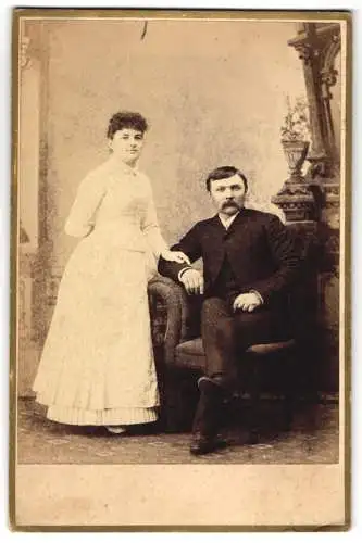 Fotografie S. M. Miller, Mason City, Dame im weissen Kleid und Herr auf Stuhl