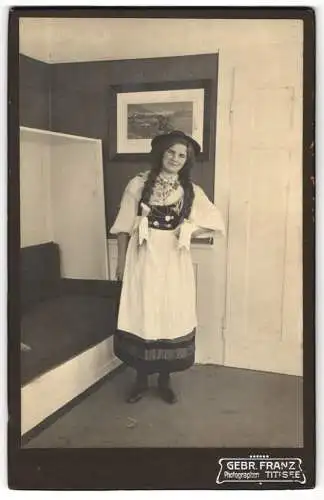 Fotografie Gebr. Franz, Titisee, junge Frau posiert in Tracht im Wohnzimmer, lange geflochtene Zöpfe