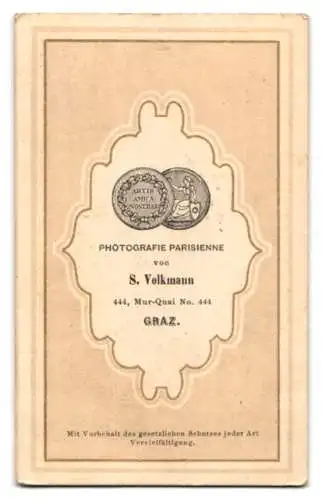 Fotografie S. Volkmann, Graz, Herr im schwarzen Wintermantel mit Vollbart stehend am Sekretär