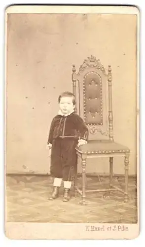 Fotografie K. Hanel et J. Plha, Ort unbekannt, niedlicher junger Knabe im Samtanzug neben einem Stuhl