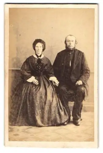 Fotografie H. Nebbin, Oldenburg, älteres Paar im dunklen weiten Kleid und im Anzug mit Backenbart