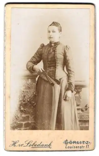 Fotografie H. v. Lobedank, Görlitz, Louisenstrasse 17, Elegante junge Dame im taillierten Kleid mit Kreuzkette u. Fächer