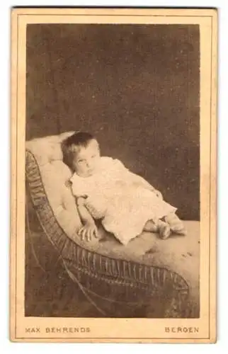 Fotografie Max Behrends, Bergen, Niedliches Kleinkind im weissen Kleid liegt ruhig auf einem Sessel