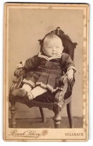 Fotografie Bernh. Schery, Sulzbach, Kleinkind im schwarzen Kleid mit weisser Strumpfhose auf einem kleinen Stuhl