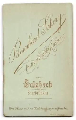 Fotografie Bernh. Schery, Sulzbach, Kleinkind im schwarzen Kleid mit weisser Strumpfhose auf einem kleinen Stuhl