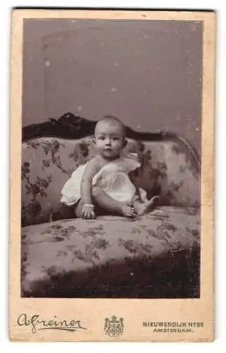 Fotografie A. Greiner, Amsterdam, Nieuwendijk 89, Kleinkind im weissen Gewand mit aufmerksamem Blick auf der Couch
