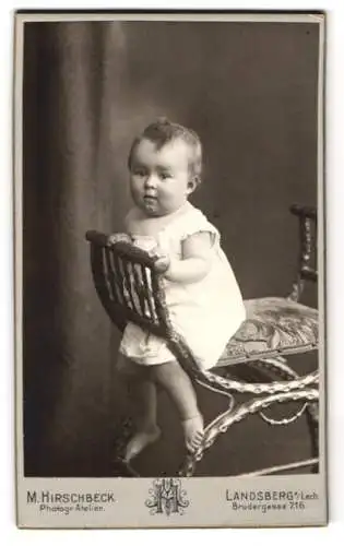 Fotografie M. Hirschbeck, Landsberg a. Lech, Brudergasse 216, Kleinkind im weissen Gewand an der Armlehne eines Stuhls